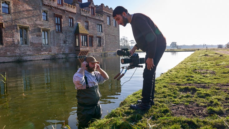 A man wearing waders stands waist-deep in water in a moat, in front of a castle. Another man on the bank of the moat is filming him with a large video camera.